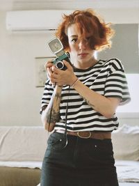 Portrait of young woman holding camera while standing at home