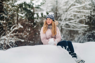 Cute girl with long hair is sitting on a mountain of snow in a hat and making snowballs
