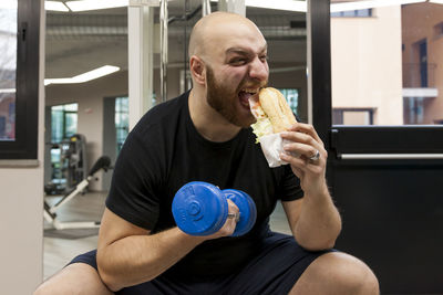 Young man eating hot dog while lifting dumbbell in gym