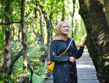 Smiling young woman standing by tree trunk in forest