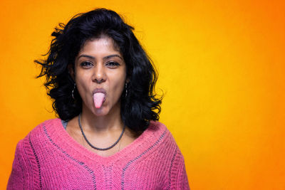 Portrait of young woman sticking out tongue against orange background