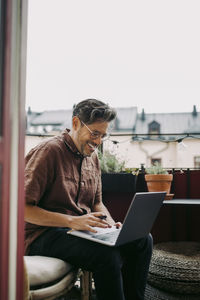 Smiling businessman working on laptop while sitting in balcony during covid-19