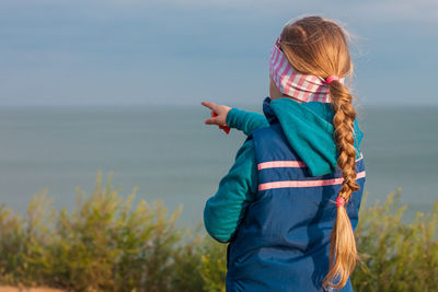 Little blond hair girl long braid on sea landscape background child staying back view  showing hand