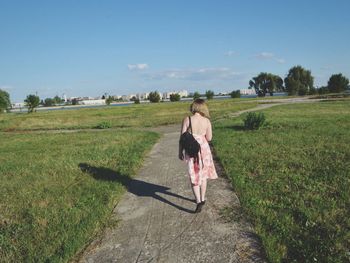 Rear view of woman walking on footpath amidst field against sky