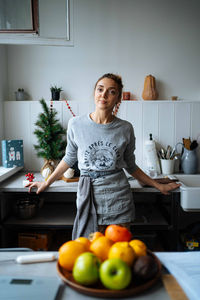 Positive adult woman in apron leaning on counter and looking at camera while cooking in kitchen at home