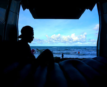 Silhouette man sitting by sea against sky seen through window