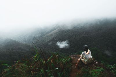 Rear view of person sitting on mountain against foggy weather