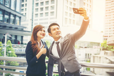 Smiling colleagues taking selfie while standing against buildings in city