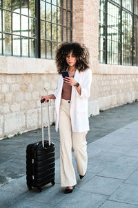 Full length hispanic female manager in stylish white suit with curly hair browsing on smartphone while walking near suitcase against old building during business trip in city