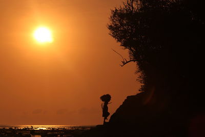 Silhouette worker carrying basket walking on rock against orange sky at shore