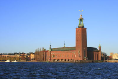 Lighthouse by sea against buildings in city against clear blue sky