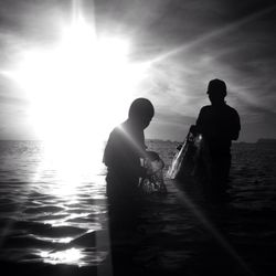 Silhouette men holding fishing nets at sea against sky on sunny day