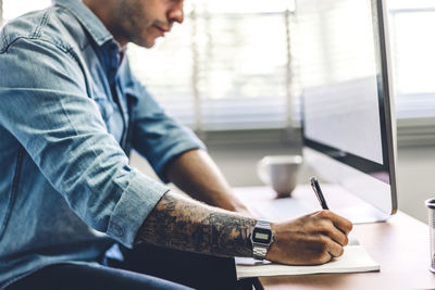 Midsection of man writing in book while using computer on table in office