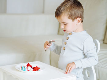 Cute toddler 3 years boy in grey pajamas eating red velvet cake in bright interior