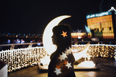 Side view of young woman standing on illuminated building terrace against clear sky at night