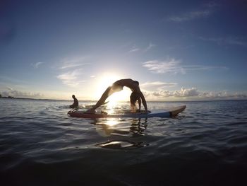 Full length of woman exercising on surfboard at lake against sky during sunset