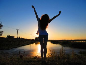 Woman with arms raised standing at lakeshore against sky during sunset