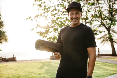 Portrait of smiling man with exercise mat standing in park