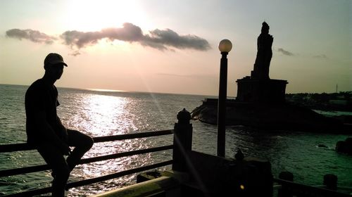 Man sitting on railing at observation point by sea against sky during sunset