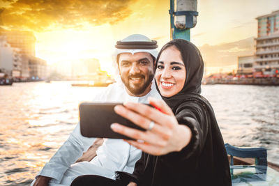 Couple talking selfie while sitting in boat on river during sunset