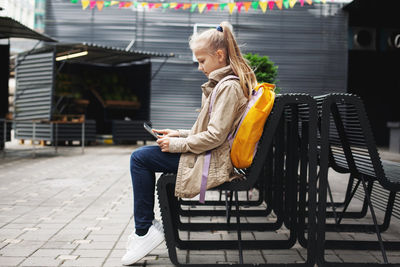 After school, a cute girl sits on a bench waiting for the bus, studies on a tablet online