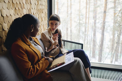 Businesswoman discussing with coworker sitting by glass window at convention center