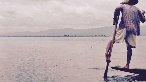 Rear view of fisherman standing on boat with oar in river against sky