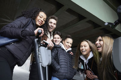 Low angle portrait of happy male and female teenage friends with electric push scooters below bridge