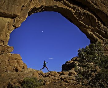 Low angle view of woman jumping on rock formation against clear blue sky