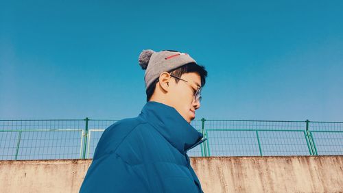 Young man looking down while standing against wall and clear blue sky