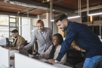 Group of diverse cheerful coworkers gathering at table with laptop discussing project while working together in contemporary workspace