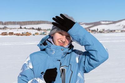 A young girl skier is blocked by a hand from the sun, stands on a frozen lake against village.