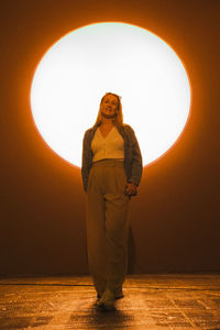 Side view of young woman standing against orange art light installation 