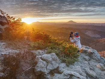 Woman hiker and photographer set tripod with camera on exposed rock above deep misty valley