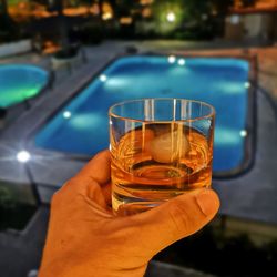 Cropped hand of man having whiskey in glass against swimming pool at night