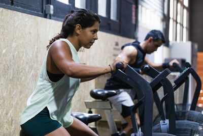 Muscular hispanic sportswoman in activewear doing exercise on air bike during workout in light gym near sportsman with equipment