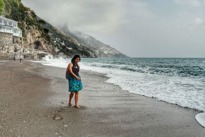 Full length of young woman standing at beach against cloudy sky