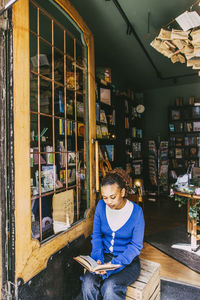 Young woman reading book while sitting near door at bookstore