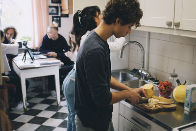 Teenage boy with female cutting fruits at kitchen counter while friends sitting in background