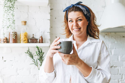 Attractive young woman plus size body positive in white shirt drink morning coffee at home kitchen