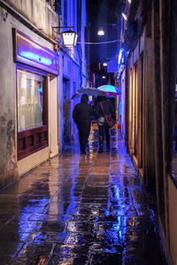 Man walking on wet illuminated street amidst buildings in city during rainy season