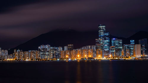 Illuminated modern buildings by bay against sky at night