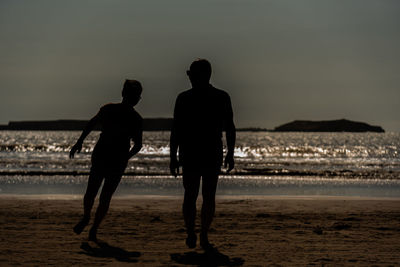 Rear view of silhouette men standing at beach against sky during sunset
