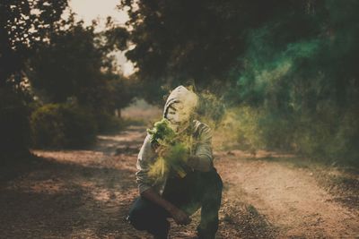 Portrait of man wearing mask while holding distress flare on dirt road amidst trees