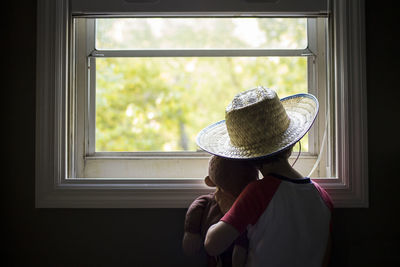 Rear view of boy holding stuffed toy while looking through window at home