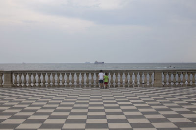 Rear view of people walking on tiled floor by sea against sky