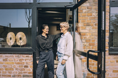 Happy female owner with hand on shoulder of colleague standing at doorway of clothing store