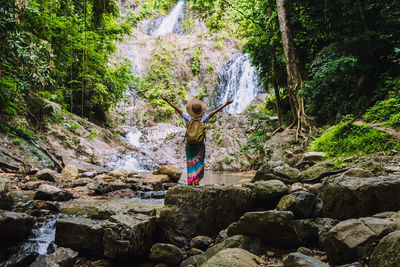 Rear view of woman with arms raised looking at waterfall in forest