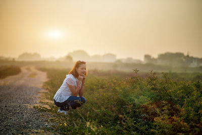 Woman crouching on field against sky during sunset