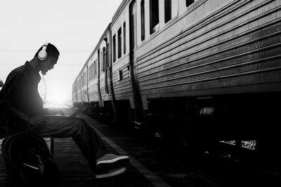 Side view of young woman sitting on railroad tracks against sky